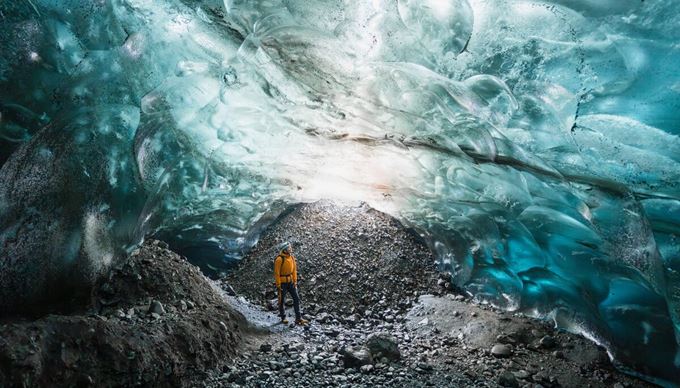 Light reflecting through blue crystal ice cave tourist in safety gear poses for photo