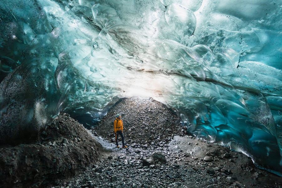 Light reflecting through blue crystal ice cave tourist in safety gear poses for photo