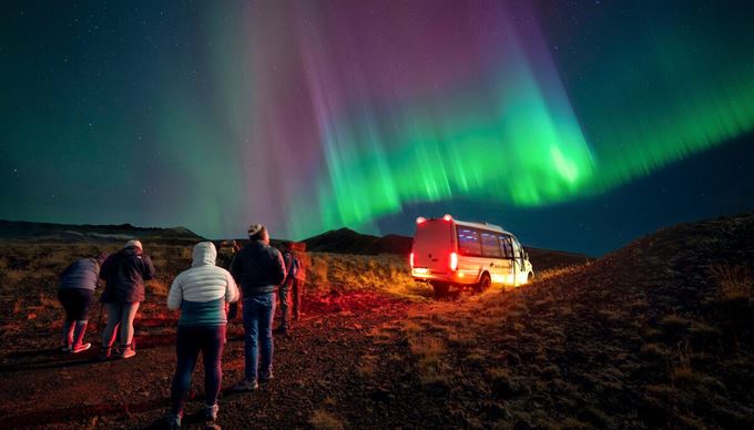 Group of tourists standing next to Arctic Adventures sprinter van underneath northern lights in South West Iceland.