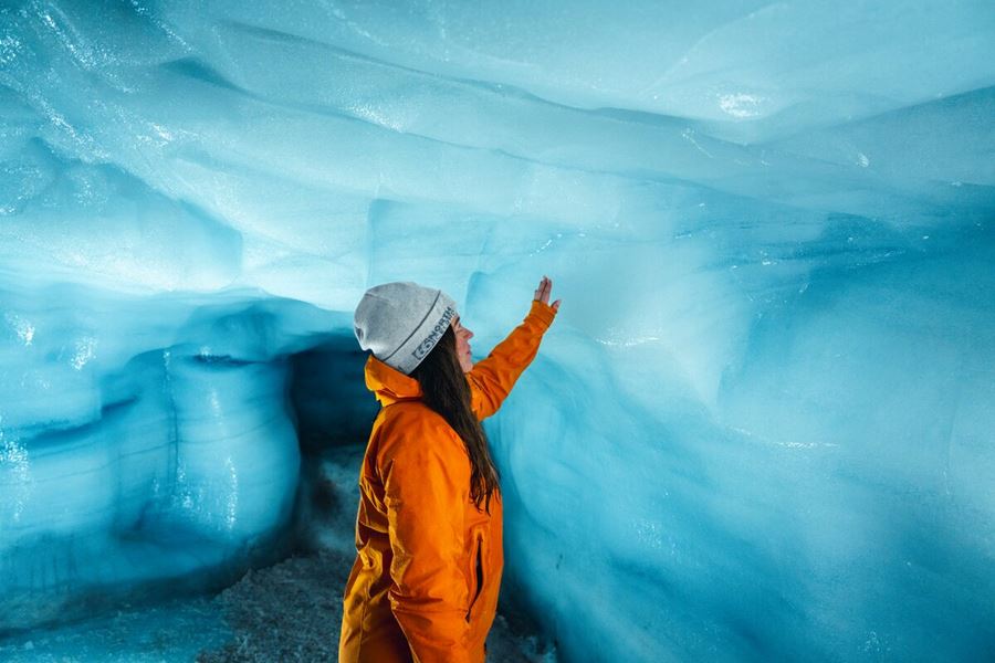 Lady in orange coat reaching to blue ice wall inside ice tunnel 